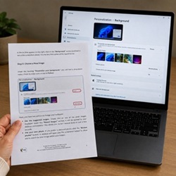 A person holding a printed sheet from PC guides for beginners and seniors next to a laptop showing the Windows background settings screen.